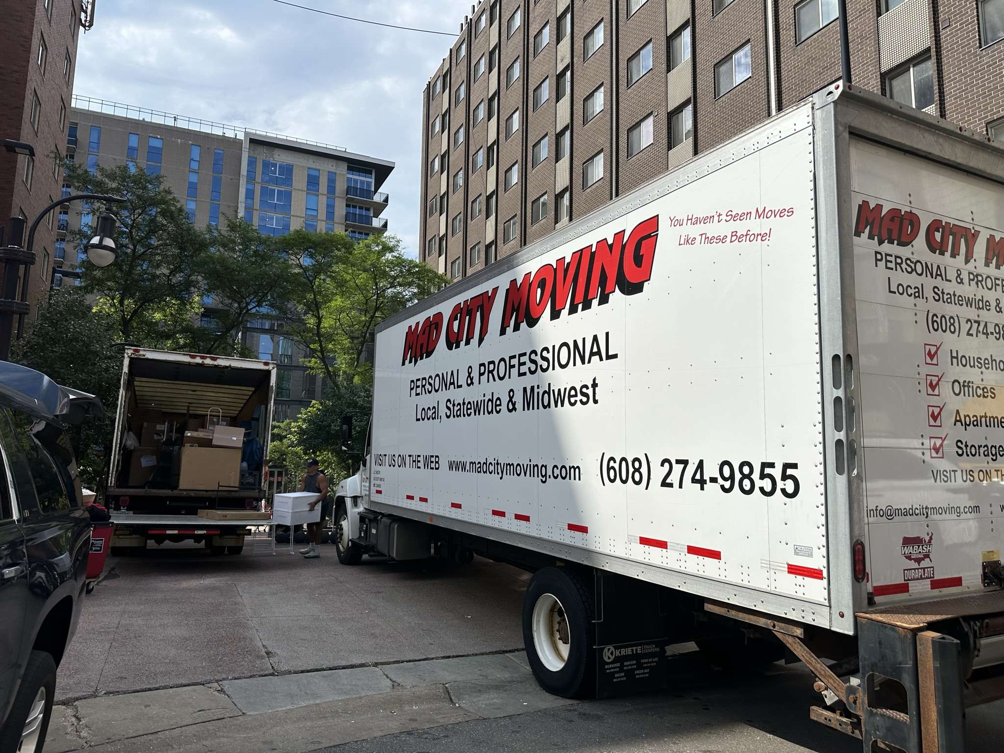 Two Mad City Moving company trucks in between buildings. One is closed showing the side promotional print stating "Mad City Moving Personal & Professional Local, Statewide & Midwest"