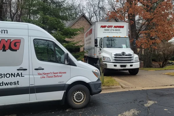 Two moving trucks are parked outside a house in a suburban neighborhood on a cloudy day. One truck is on the street, and the other is backed into a driveway. Trees and houses are visible in the background.