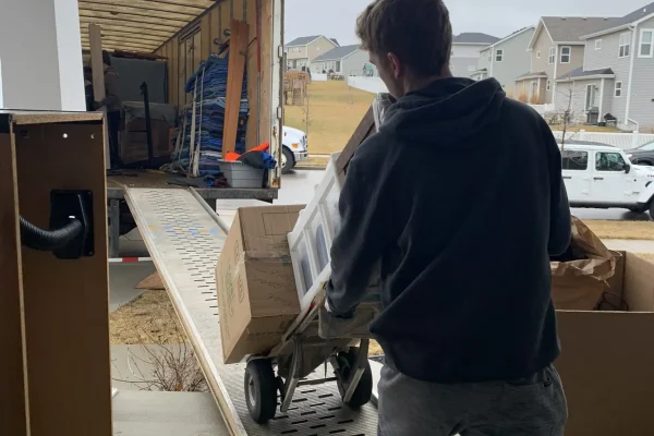 A person in a hoodie and boots pushes a dolly loaded with boxes up a ramp into a moving truck from the best moving company in Madison, parked outside a house in a suburban neighborhood on a cloudy day.